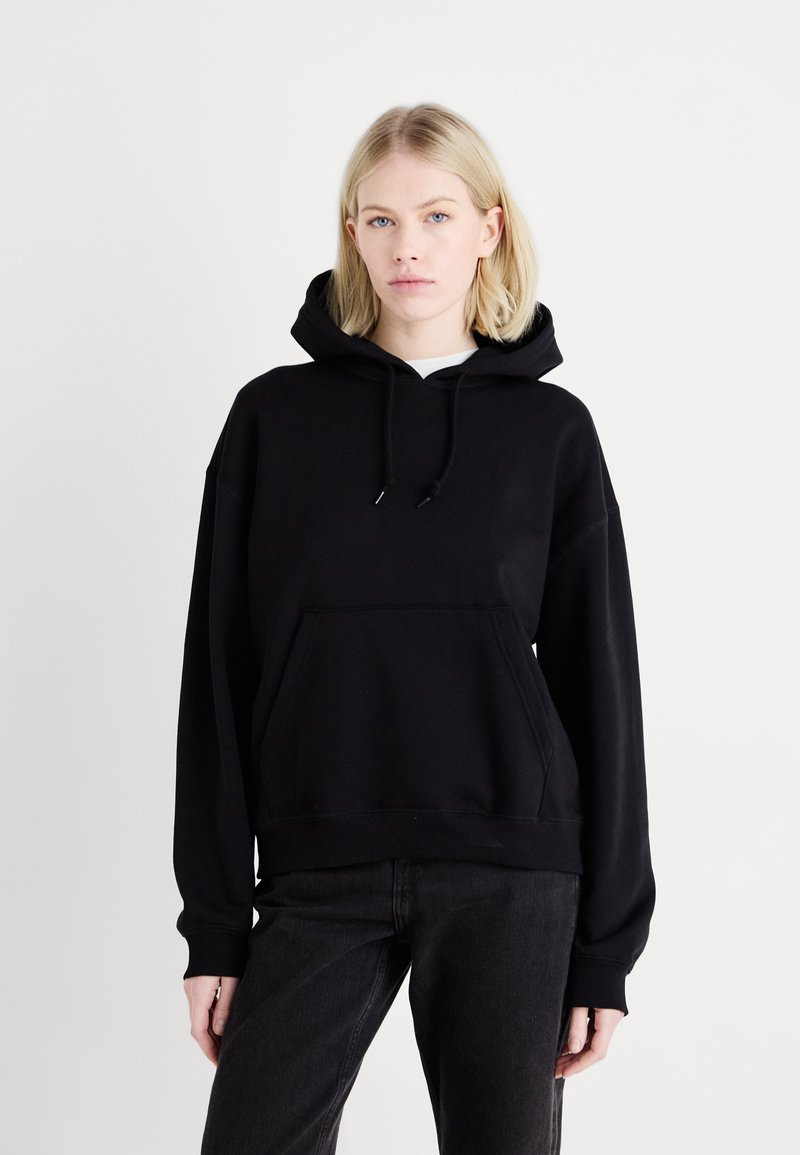 Felpa nera oversize con tasca frontale, coulisse nel collo e polsini e orlo a costine. Indossata sopra una maglietta bianca e abbinata a pantaloni neri.