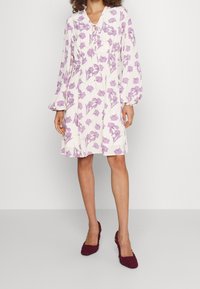 Helles rosa Kleid mit lila floralem Muster, langen Ärmeln und einer Schleifenhalslinie. Der Stoff wirkt weich und fließend und reicht bis zur Mitte des Oberschenkels.