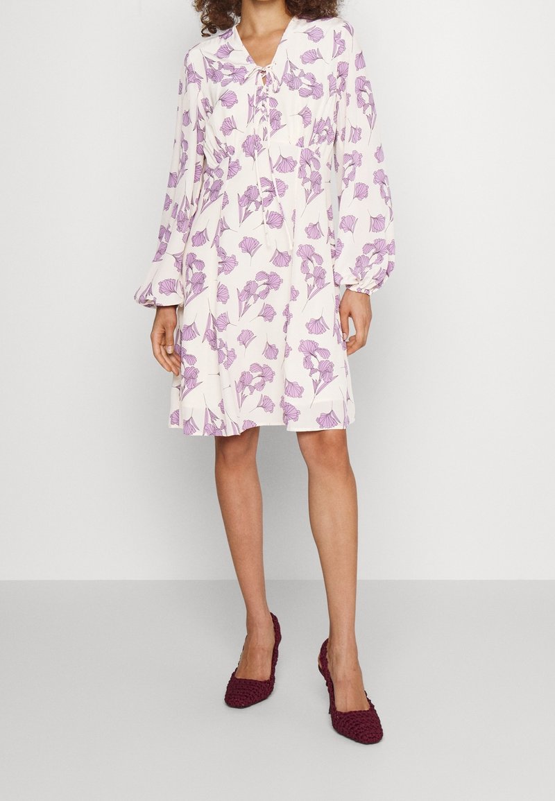 Helles rosa Kleid mit lila floralem Muster, langen Ärmeln und einer Schleifenhalslinie. Der Stoff wirkt weich und fließend und reicht bis zur Mitte des Oberschenkels.