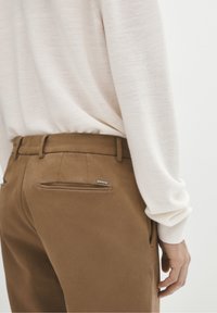 Osoba w beżowych spodniach i kremowej bluzce z długimi rękawami, z jedną ręką swobodnie opartą przy boku na jednolitym tle.