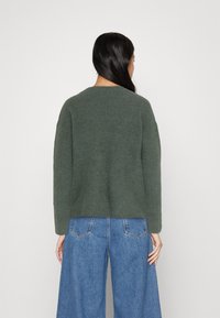 Maglione verde a coste con scollo rotondo e spalle cadenti, abbinato a jeans a gamba larga azzurri. La texture sembra morbida e accogliente.