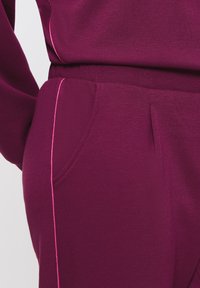 Vinröd träningsdräkt med en mjuk textur, med rosa detaljer längs sömmarna och en sidoficka. Tyget verkar sträckt och slätt.