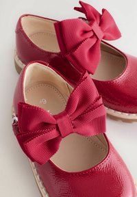 Scarpe di vernice rossa con grandi fiocchi texture, punte arrotondate e suola leggera. L'interno ha una fodera beige liscia.