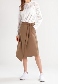 Top en maille blanche avec un motif ajouré, associé à une jupe enveloppante marron avec un nœud sur le devant. Le modèle porte des baskets blanches.