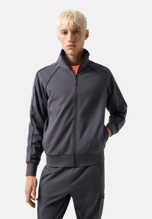 Junge Person mit kurzen blonden Haaren, bekleidet mit einer dunkelgrauen Trainingsjacke mit Reißverschluss und passenden Hose, steht mit einer Hand in der Tasche.