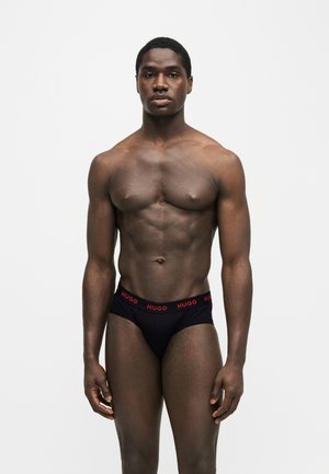 Homme musclé debout sur un fond uni, portant un slip noir avec une ceinture rouge avec le logo "HUGO".