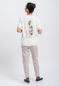 Witte katoenen t-shirt met een grafisch ontwerp op de achterkant, gecombineerd met lichtbeige broek en zwarte sneakers. Eenvoudige, casual outfit.