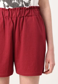 Shorts en coton rouge avec une taille élastique, présentant des détails plissés en haut et une coupe ample. Texture lisse et bords ourlés.