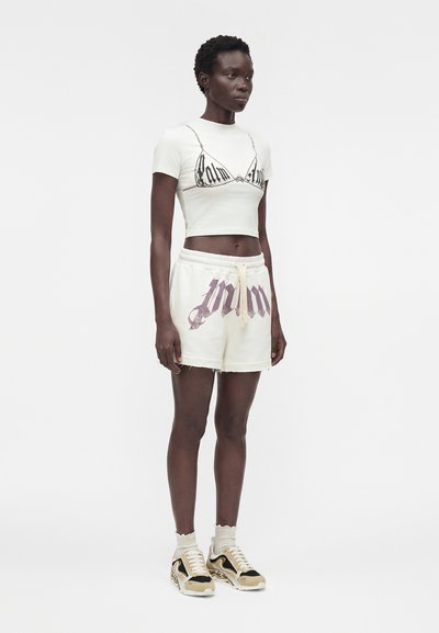 Γυναίκα στέκεται φορώντας ένα κοντό λευκό T-shirt με τύπωμα σουτιέν, λευκά σορτς με γραφικό κείμενο, μπεζ κάλτσες και μπεζ-μαύρα αθλητικά παπούτσια.