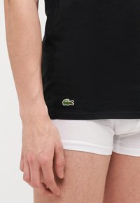 T-shirt nera in cotone a maniche corte con un logo verde a forma di coccodrillo sull'orlo, abbinata a pantaloni corti bianchi aderenti.