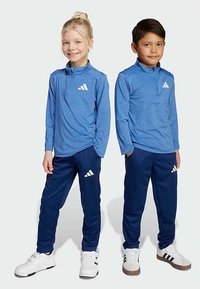 Siniset urheilupaidat neljäsosavetoketjulla, yhdistetty laivastonsinisiin verkkarihousuihin. Molemmissa valkoiset Adidas-logot. Sileä, hengittävä kangas.