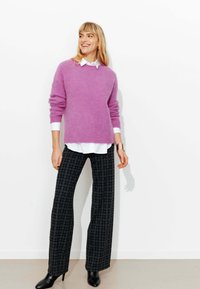 Femme portant un pull violet sur une chemise blanche, un pantalon large à carreaux noirs, des bottines à talons noires et un bracelet manchette doré, souriante et debout.