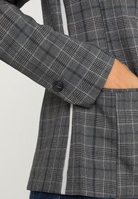 Šedý kostkovaný blazer s hladkou texturou, černým zapínáním na knoflík, bílými akcenty podél rukávu a kapsou.
