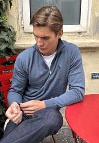 Jaunas vyras mėlynėje megztinėje su užtrauktuku sėdi prie raudono lauko stalo ir kėdės, žiūrėdamas žemyn ir reguliuodamas rankovę šalia prie išvytų vijoklių uždengtos sienos.