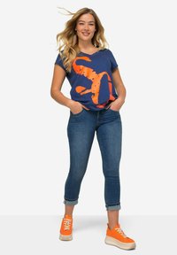 T-paita, jossa on merensinisen V-kaula-aukko ja suuri oranssi graafinen kuvasto, yhdistetty tiukkoihin sinisiin farkkuihin ja oransseihin suosikkitossuihin, joissa on valkoiset pohjat.