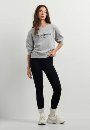 Jonge vrouw staat met hand in de taille, draagt een grijze Pepe Jeans trui, zwarte legging, witte sokken en beige sneakers tegen een effen achtergrond.