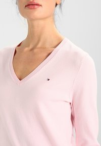 Jasnoróżowy sweter z dekoltem w serek, z ryżowym wykończeniem, miękką teksturą i małym haftowanym logo na klatce piersiowej.