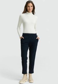 Weißer, gerippter Rollkragenpullover kombiniert mit dunkelblauen Cordhosen mit Seitentaschen, ergänzt durch beige Stiefeletten.