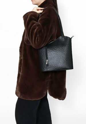 Sac fourre-tout noir texturé avec une fermeture éclair et double poignées, arborant un motif à points en relief. Porté avec un manteau en fausse fourrure marron.