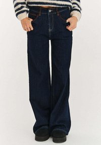 Tmavé džínové kalhoty s širokými nohavicemi a vysokým pasem, zdobené hnělým šitím, předními kapsami a klasickým zapínáním na knoflíky a zip.