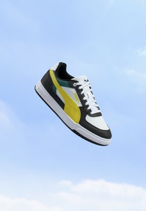 Baskets Puma blanches, noires et jaunes avec des lacets blancs flottant sur un fond de ciel bleu.