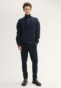 Marineblauer Strickpullover mit einem Halbreißverschluss-Kragen, der ein strukturiertes Muster und gerippte Ärmel aufweist. Kombiniert mit dunklen Hosen und schwarzen Stiefeln.
