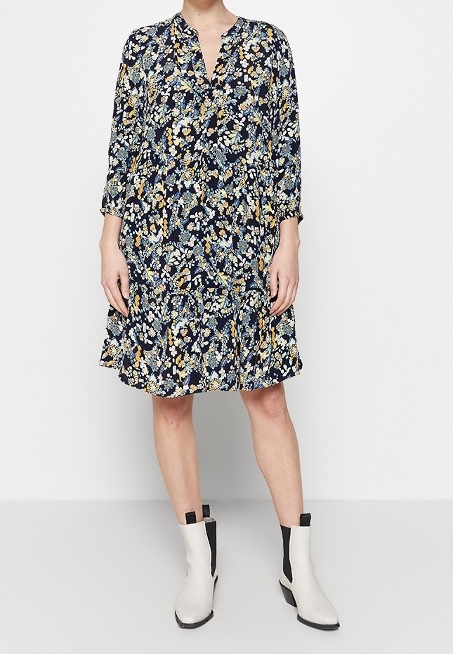 Robe fleurie bleu marine à manches longues, avec un décolleté en V et un ourlet à volants. Assortie à des bottines blanches avec des détails noirs.