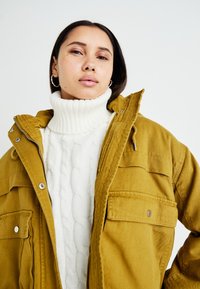 Geel corduroy jack met meerdere voorkruiszakken, gelaagd over een wit kabelbrei turtlenecktrui. Soepele textuur en casual ontwerp.