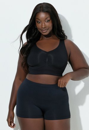 Kobieta plus size z długimi włosami, ubrana w czarny sportowy biustonosz i czarne szorty, pozująca na białym tle.