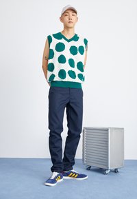 Pulover fără mâneci verde și alb cu buline, pantaloni bleumarin și adidași albaștri cu dungi galbene; cutie de metal argintie de depozitare în apropiere.