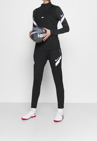 Czarny długi rękaw sportowy top z półzamkiem, w połączeniu z dopasowanymi czarnymi spodniami z białymi akcentami oraz białymi butami sportowymi z czerwonymi podeszwami. Trzyma czarną piłkę nożną.