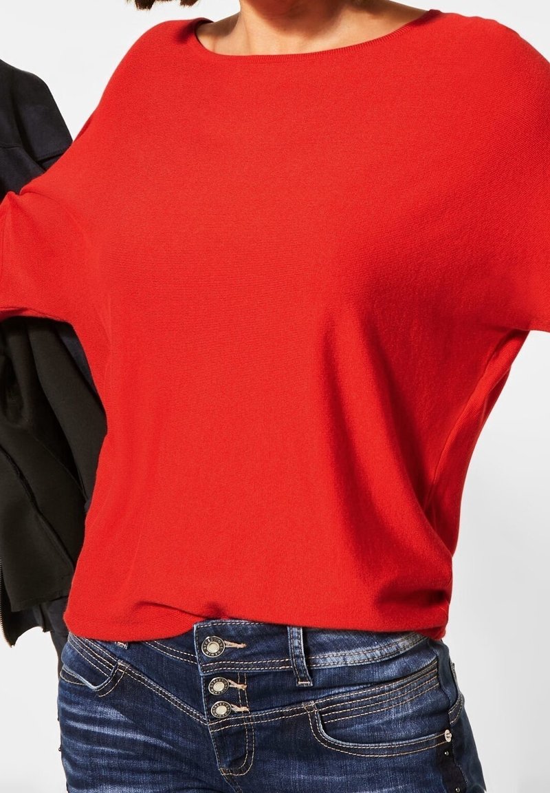 Red knit pullover with a wide neckline and dropped shoulders, paired with dark blue jeans featuring decorative metal buttons.