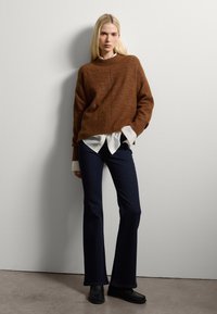 Un pull en tricot marron avec un ourlet côtelé porté sur une chemise blanche, associé à un jean évasé noir et des chaussures noires, sur un fond neutre.