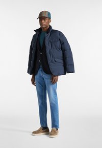 Giacca a vento blu navy con colletto alto, jeans azzurri chiari e scarpe slip-on color beige. Un cappello a fantasia completa l'outfit con un contrasto di texture.