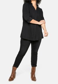 Frau in einem lockeren schwarzen Shirt, engen schwarzen Hosen und braunen, hochhackigen Stiefeletten, die mit einem Arm den anderen überkreuzt dasteht.