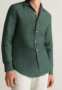 Camisa de lino verde oscuro con una textura suave, mangas largas, cuello puntiagudo y botones en la parte delantera, con dobladillo curvado.