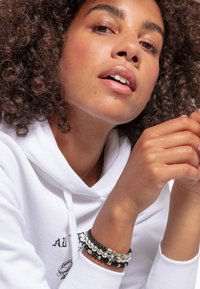 Sudadera blanca con estampado gráfico negro. Varios brazaletes de cuentas en materiales negros, plateados y transparentes en la muñeca. Cabello rizado enmarcando el rostro.