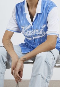Maillot rayé bleu à manches blanches, affichant un logo "Kani" en écriture cursive. Le modèle porte un jean en denim clair avec un tatouage visible sur son bras.