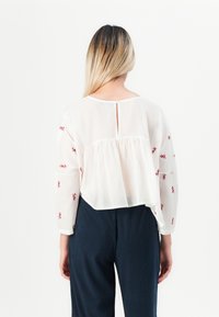 Woman with straight blonde hair wearing a loose white blouse embroidered with red flowers and dark blue pants, standing against a white background.