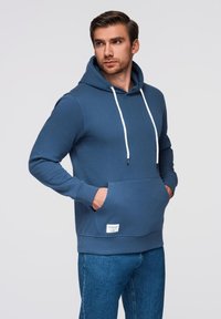 Felpa blu realizzata in tessuto morbido con tasca a marsupio, cordini bianchi e un piccolo etichetta all'orlo. Indossata con jeans blu.