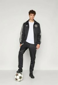 Svart idrettsdrakt med hvite striper, bestående av en jakke med glidelås og matchende bukser. Modellen står på en svart og hvit fotball.