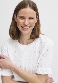 Hvid broderet skjorte med korte, voluminøse ærmer, der har et struktureret mønster og små udsnit i stoffet.