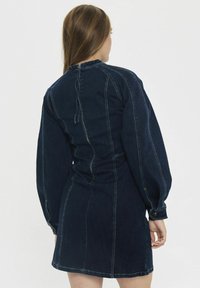 Sötét denim ruha hosszú, puffos ujjakkal, karcsú derékkal és cipzáros záródással a hátsó részen. Kontrasztos varrási részletek az oldalvarrások mentén.