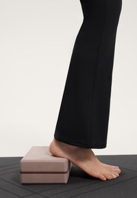 Zwarte wijde broek bedekt een deel van een voet die rust op een lichtroze rechthoekig blok met een horizontale bruine streep. Gelaagd donkere oppervlakte daaronder.