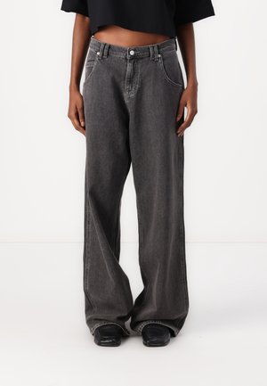 Personne portant un jean noir délavé et ample ainsi que des chaussures noires à bout carré sur un fond blanc uni.