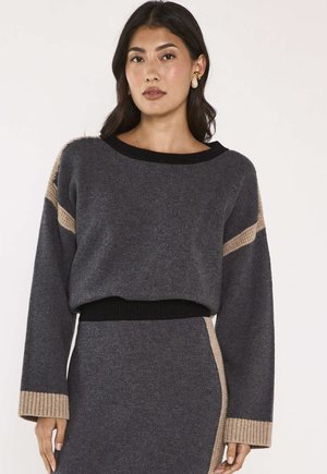 Donna che indossa un maglione di lana grigio scuro e gonna abbinata con accenti beige, in piedi davanti a uno sfondo chiaro e semplice.