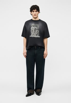 Ung mand med krøllet hår iført en sort grafisk t-shirt med teksten "NON HUNDRED", brede sorte jeans og spidse sorte sko.
