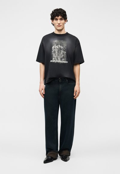 Young man with curly hair wearing a black graphic t-shirt reading "NON HUNDRED," wide black jeans, and pointed black shoes.