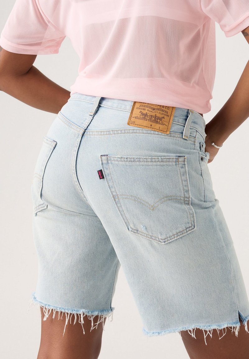 Helle blaue Jeansshorts mit ausgefransten Säumen, ausgestattet mit einem Leder-Patch auf der Rückseite und unverwechselbarer Levi's-Markierung auf der Gesäßtasche.
