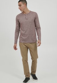 Langærmet, maroon henley skjorte med tre knapper ved halsen, parret med beige bukser og sorte sneakers. Glat stoftekstur.